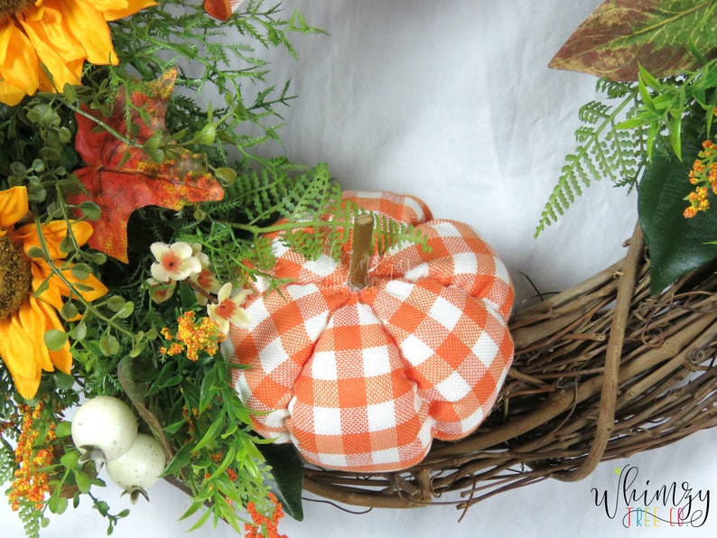 Pode incluir: Uma coroa de outono com base de videira marrom, decorada com girass&oacute;is, folhas verdes, flores brancas e uma pequena ab&oacute;bora xadrez laranja e branca.