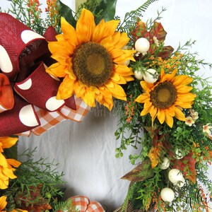 Pode incluir: Uma coroa de outono com um la&ccedil;o xadrez vermelho e branco, dois girass&oacute;is e bagas brancas. A coroa &eacute; feita de vegeta&ccedil;&atilde;o e tem um toque r&uacute;stico.