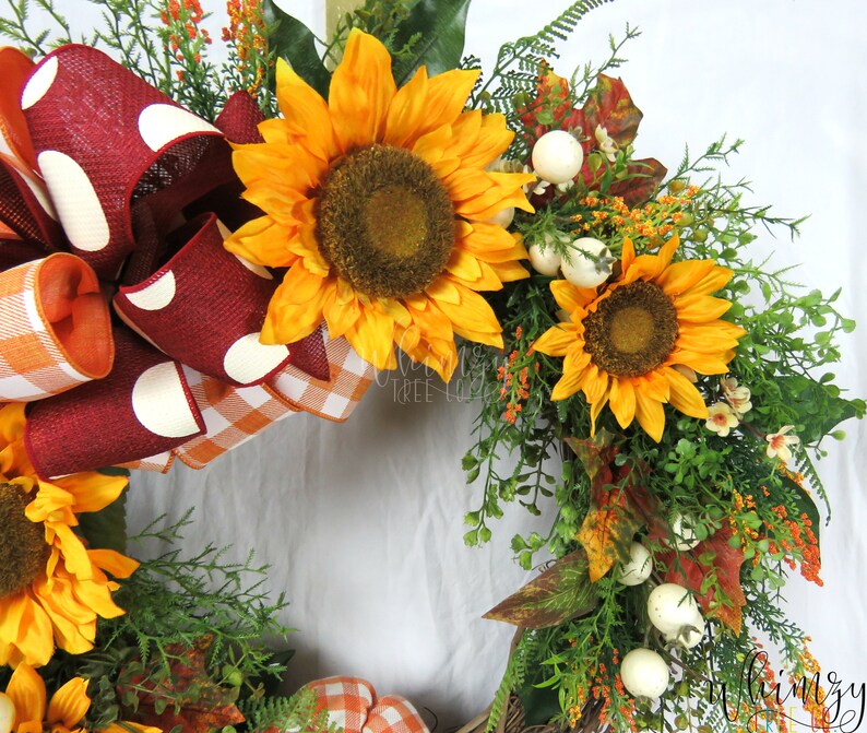 Pode incluir: Uma coroa de outono com um la&ccedil;o xadrez vermelho e branco, dois girass&oacute;is e bagas brancas. A coroa &eacute; feita de vegeta&ccedil;&atilde;o e tem um toque r&uacute;stico.