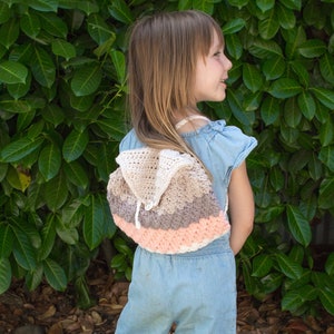 May include: A young person wearing a light blue jumpsuit with a crocheted backpack in shades of pink, white, and gray. The backpack has a hood.