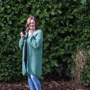 May include: A woman wearing a teal green crocheted cardigan with long sleeves and a relaxed fit. She is standing in front of a green leafy background.