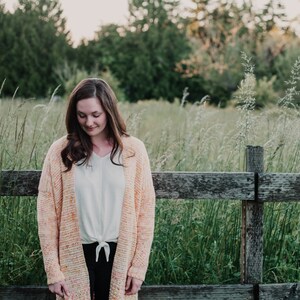 May include: A woman wearing a light pink and orange crochet cardigan with a white t-shirt tied at the waist and black trousers. She is standing in front of a wooden fence with tall grass behind her.