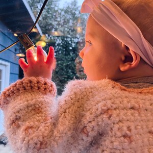 Può includere: Una bambina con un maglione rosa lavorato a uncinetto e una fascia bianca guarda fuori da una finestra. La finestra è decorata con luci a catena.