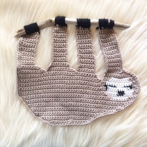 May include: A crocheted sloth hanging from a branch. The sloth is brown with a white face and black eyes.