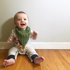 May include: A baby boy wearing a green crocheted bib and a beige onesie with a yellow bow tie. He is sitting on a wooden floor and smiling.