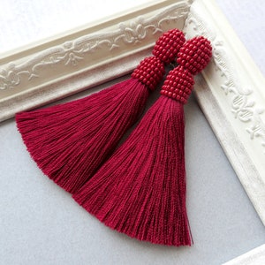 May include: A pair of long, burgundy tassel earrings with red bead accents. The earrings are displayed on a white frame.