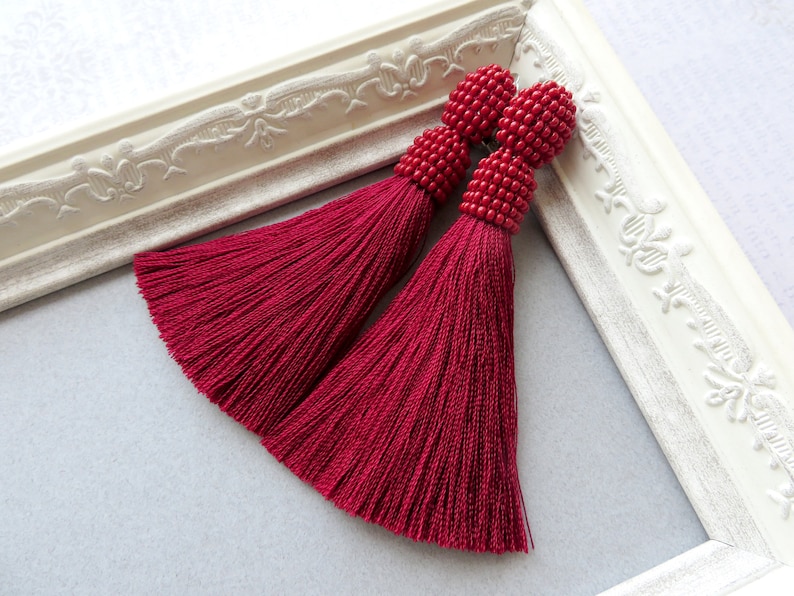 May include: A pair of long, burgundy tassel earrings with red bead accents. The earrings are displayed on a white frame.
