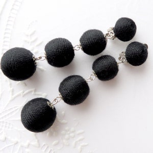 Peut inclure: Une paire de boucles d'oreilles noires à clip. Chaque boucle d'oreille est composée d'une série de sphères noires recouvertes de tissu, reliées par des chaînes argentées. Les boucles d'oreilles sont présentées sur une surface blanche texturée.