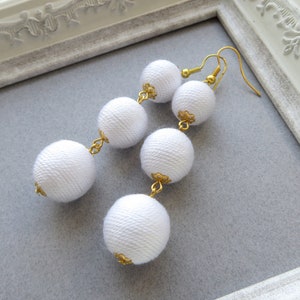 Op de afbeelding: Lange witte stoffen bal oorbellen met gouden accenten.