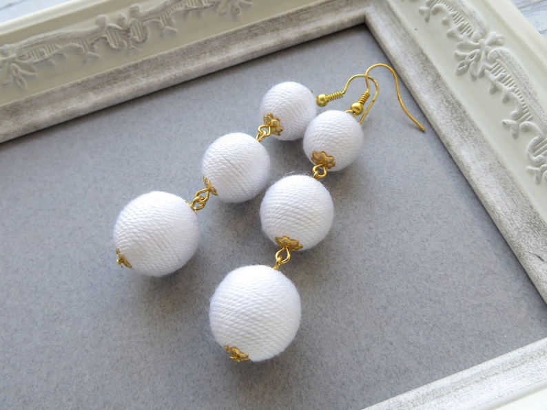 Op de afbeelding: Lange witte stoffen bal oorbellen met gouden accenten.