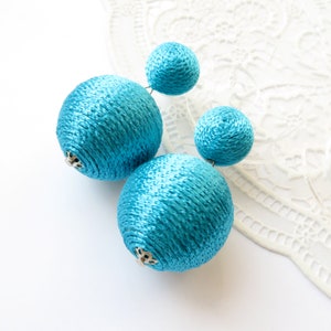 May include: Turquoise blue statement earrings with a textured, woven appearance. The earrings feature four spherical shapes, two larger and two smaller, connected by thin wires. The earrings are set against a white lace background.