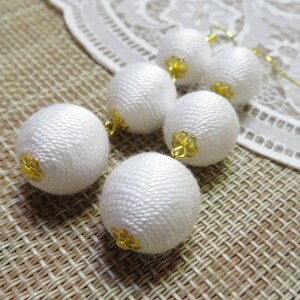 Op de afbeelding: Witte stoffen bal oorbellen met gouden accenten. De oorbellen hangen en hebben meerdere stoffen ballen.