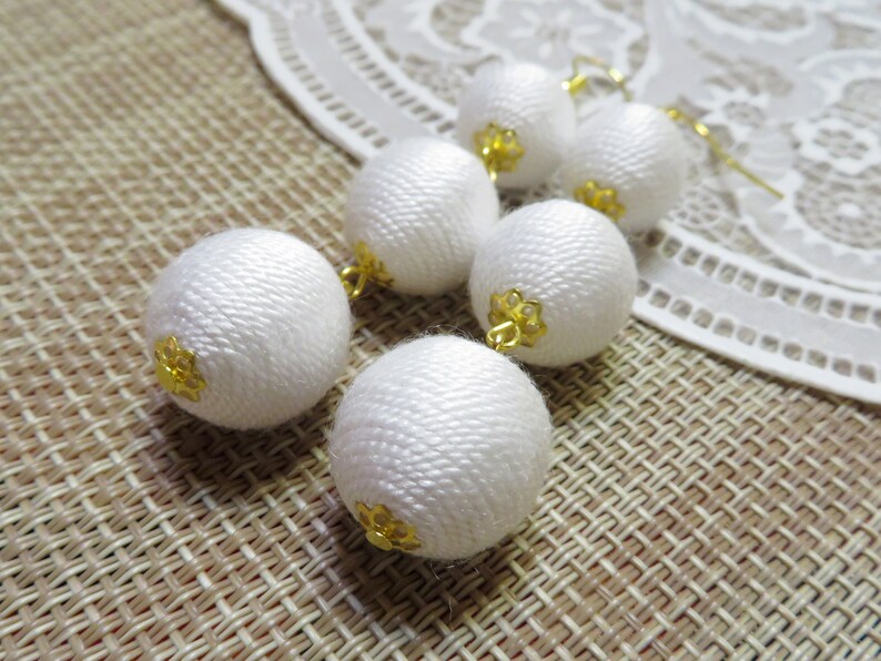 Op de afbeelding: Witte stoffen bal oorbellen met gouden accenten. De oorbellen hangen en hebben meerdere stoffen ballen.