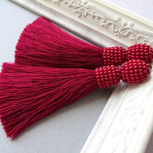 May include: A pair of long, burgundy tassel earrings with red bead accents. The tassels are made of a soft, silky material and the beads are round and evenly spaced.