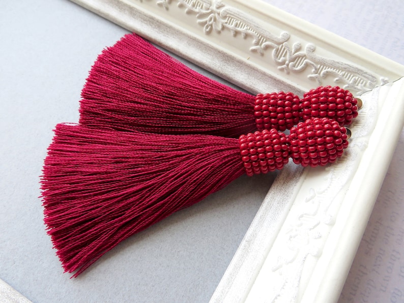 May include: A pair of long, burgundy tassel earrings with red bead accents. The tassels are made of a soft, silky material and the beads are round and evenly spaced.