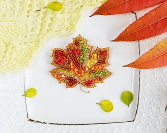 May include: A beaded maple leaf brooch in shades of red, orange, yellow, and green. The brooch is on a white plate with fall leaves and a knitted fabric.
