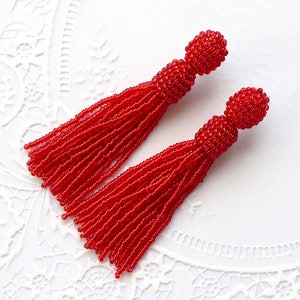 Può includere: Un paio di orecchini a nappina di perline rosse. Gli orecchini sono realizzati con piccole perline rotonde che sono infilate insieme per creare una forma a nappina. Le nappine sono lunghe circa 5 cm e gli orecchini circa 7,5 cm.