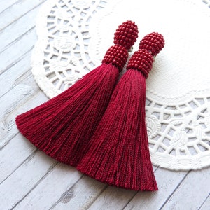 May include: A pair of long, red tassel earrings with beaded tops. The tassels are made of a soft, silky material and hang down to about 3 inches in length.