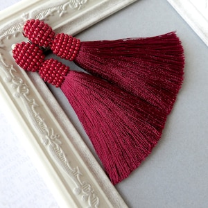 May include: A pair of burgundy tassel earrings with red beaded tops. The earrings are displayed against a white background.