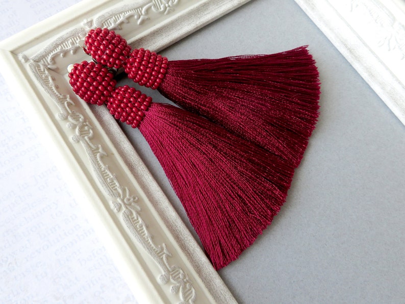 May include: A pair of burgundy tassel earrings with red beaded tops. The earrings are displayed against a white background.