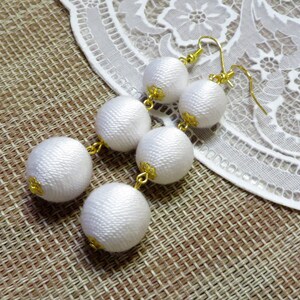 Op de afbeelding: Witte zijden bal oorbellen met gouden accenten. De oorbellen hangen en hebben drie ballen aan elke oorbel.