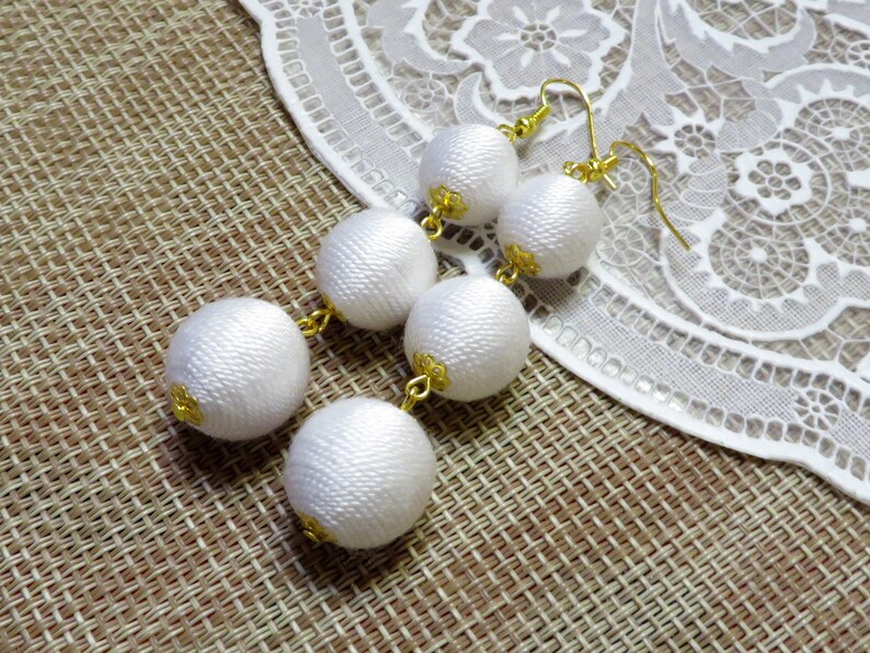 Op de afbeelding: Witte zijden bal oorbellen met gouden accenten. De oorbellen hangen en hebben drie ballen aan elke oorbel.