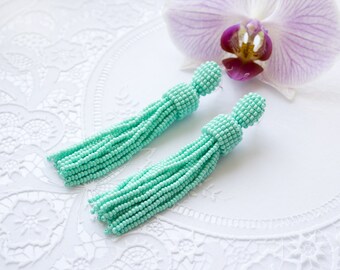 May include: A pair of mint green beaded tassel earrings. The earrings are made of small, round beads that are strung together to create a tassel shape. The tassels are approximately 7.5 cm long and are attached to a small, round bead at the top.