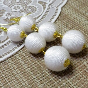 Op de afbeelding: Witte stoffen bal oorbellen met gouden accenten. De oorbellen hangen en hebben meerdere ballen aan elke kant.