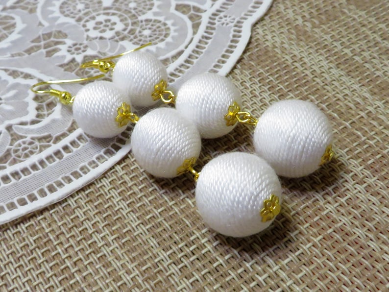 Op de afbeelding: Witte stoffen bal oorbellen met gouden accenten. De oorbellen hangen en hebben meerdere ballen aan elke kant.