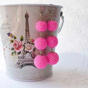 Puede incluir: Pendientes de gota de cuentas rosas con tres cuentas rosas en cada pendiente. Los pendientes cuelgan de un cubo blanco con un diseño de la Torre Eiffel parisina.
