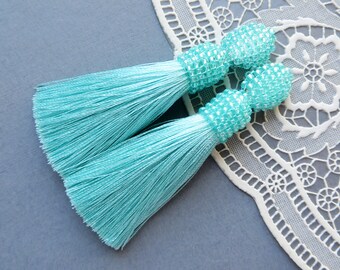 May include: A pair of turquoise blue tassel earrings with beaded tops. The earrings are on a white lace background.