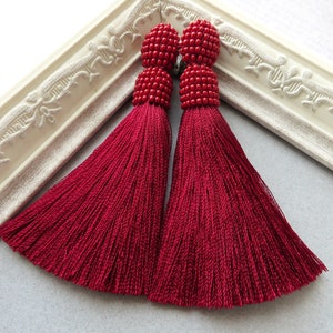 May include: A pair of long, red tassel earrings with beaded tops. The tassels are made of a soft, silky material and hang down to about 3 inches in length.