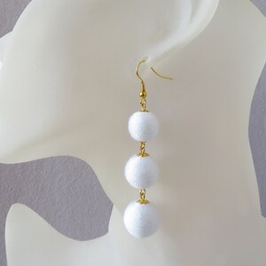 Op de afbeelding: Witte stoffen pompon oorbellen met een gouden ketting en haak. De oorbellen hangen aan het oor van een wit mannequin.