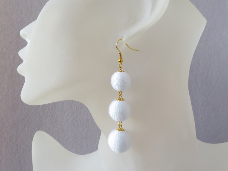 Op de afbeelding: Witte stoffen pompon oorbellen met een gouden ketting en haak. De oorbellen hangen aan het oor van een wit mannequin.
