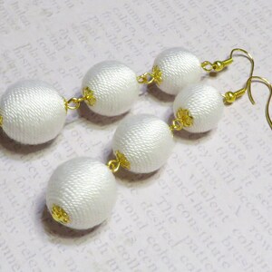 Op de afbeelding: Een paar witte stoffen oorbellen met gouden accenten. Elke oorbel heeft drie ronde stoffen ballen die met elkaar verbonden zijn door gouden kettingen.