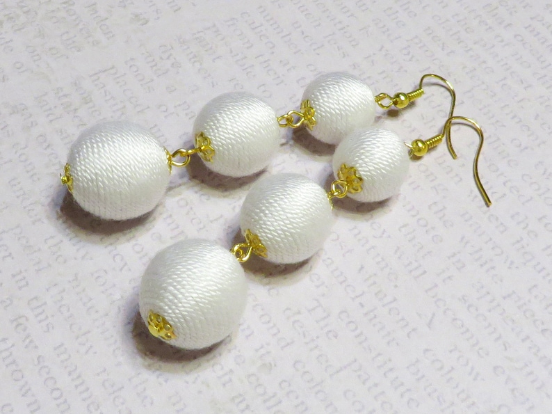 Op de afbeelding: Een paar witte stoffen oorbellen met gouden accenten. Elke oorbel heeft drie ronde stoffen ballen die met elkaar verbonden zijn door gouden kettingen.