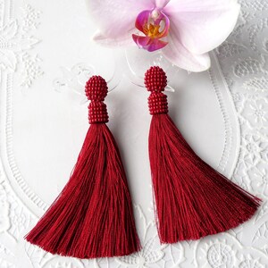 Peut inclure: Une paire de longues boucles d'oreilles à pompon rouge avec des dessus perlés. Les boucles d'oreilles sont sur un fond blanc avec un motif de dentelle blanche.