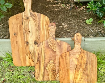 May include: Three wooden cutting boards of varying sizes, made from olive wood, with a natural wood grain pattern. The boards have a handle on the top and are arranged in a row on a bed of green grass.