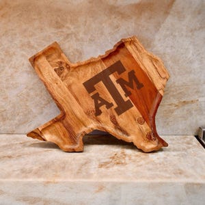 Puede incluir: Una bandeja de madera con la forma del estado de Texas, con un acabado de veta de madera natural y las letras ATM grabadas en marrón oscuro en el centro. La bandeja tiene una superficie tallada y hundida con bordes irregulares y orgánicos.
