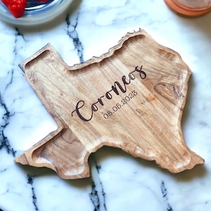 Puede incluir: Una bandeja de madera de color marrón claro con la forma del estado de Texas. La bandeja presenta un patrón de veta de madera natural y está grabada con el texto "Coroneos" en letra cursiva, seguido de la fecha "08.05.2023" en fuente simple.