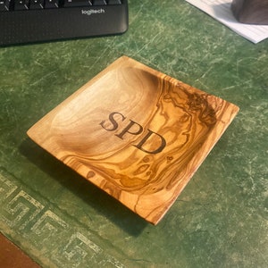 May include: A square wooden tray with a natural wood grain finish. The tray has a shallow bowl shape and is engraved with the initials "SPD".
