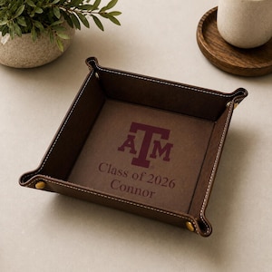 May include: A brown leather tray with white stitching and gold-colored rivets at the corners. The tray features the letters "A" and "M" in maroon, along with the text "Class of 2026 Connor".