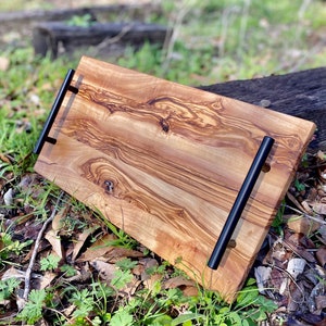 May include: A long, rectangular wooden serving tray with a natural wood grain pattern and black metal handles. The tray is sitting on a bed of green leaves and brown twigs.