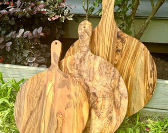 May include: Three round wooden serving boards with handles, crafted from olive wood. Each board features a distinct natural grain pattern with swirling light and dark brown wood tones. The boards are arranged in a tiered stack, showing varying sizes.