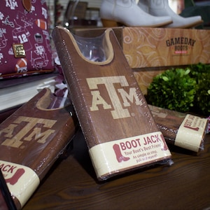 May include: Wooden boot jacks with the letters "A&M" are displayed. The boot jacks have a light brown finish and are wrapped in clear plastic. The text "Boot Jack" is printed on a white label. A maroon handbag and other items are in the background.