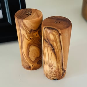 May include: Two wooden salt and pepper shakers made from olive wood. The shakers are cylindrical and have a natural wood grain pattern.