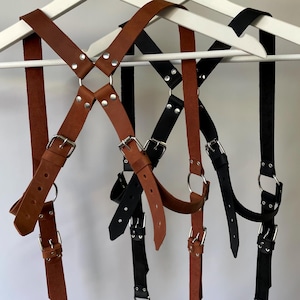 May include: Two leather body harnesses hanging on white hangers. One harness is brown and the other is black. Both feature adjustable straps with silver-toned metal buckles, rivets, and circular rings connecting the intersecting leather straps.