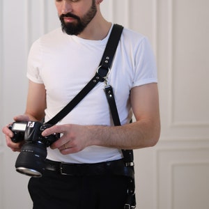 May include: A man wearing a white t-shirt and black pants is holding a black DSLR camera with a lens attached. He is wearing a black leather camera strap with a silver buckle.