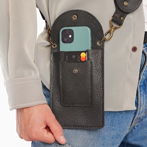 May include: A black leather phone holster with a strap and a pocket for cards. The phone holster is worn on a person's hip. The phone is light blue and has a black case. The phone holster is made in Ukraine.
