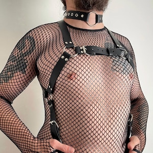 May include: A black long-sleeve fishnet shirt worn under a black leather chest harness with silver-toned metal hardware, including buckles and O-rings. A matching black leather choker with a central silver-toned O-ring is worn around the neck.